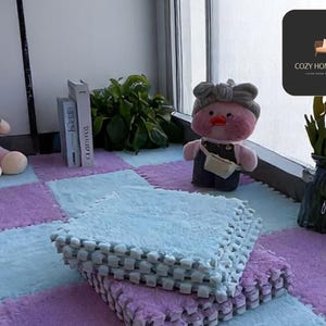 May include: A cozy indoor space featuring interlocking foam floor mats in light blue, lavender, and pink. Plush toys, including a monkey and a duck, are placed on the mats. A vase of yellow flowers and decorative books complete the scene.