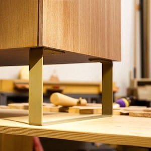 Puede incluir: Primer plano de un mueble de madera con acabado marrón claro. El mueble está sostenido por patas de metal dorado. Las patas rectangulares están unidas a la base del mueble. El mueble está sobre una superficie de madera.