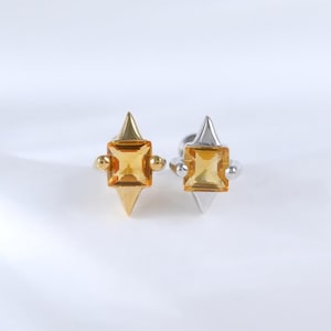 Op de afbeelding: Twee ringen met gele vierkante edelstenen. Eén ring is goudkleurig, de andere zilverkleurig. Beide hebben een driehoekige basis. De ringen worden getoond tegen een witte achtergrond.