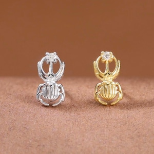 May include: Two stud earrings shaped like scarab beetles, one in silver tone and one in gold tone. Each earring features a small, clear, round-cut gemstone set at the top of the beetle design, with detailed textured lines on the body.