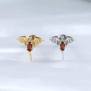 May include: Two dragon-shaped stud earrings. One is gold-toned, the other silver-toned. Each has a red gemstone body and outstretched wings. The earrings are displayed on a white surface, showcasing their intricate design and color contrast.