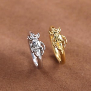 Può includere: Due anelli, uno color argento e uno color oro, ciascuno caratterizzato da un dettaglio a forma di scarabeo sulla fascia. Gli anelli sono posizionati fianco a fianco, mettendo in risalto la finitura metallica e il design scolpito dell'insetto.