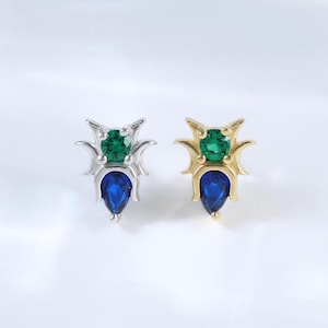 May include: A pair of insect-shaped stud earrings. One earring is silver-toned, the other gold-toned. Each features a green gemstone head and a blue teardrop-shaped gemstone body. The earrings are set against a white background.