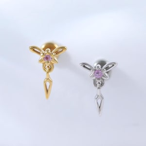 Puede incluir: Dos pendientes de botón, uno dorado y otro plateado, cada uno con un diseño de abeja, una gema rosa y un dije colgante en forma de lágrima. Los pendientes se muestran sobre una superficie blanca.