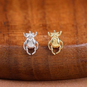 May include: Two small beetle-shaped stud earrings, one in a polished silver tone and the other in a polished gold tone. Each earring features a detailed insect body with visible legs and antennae, designed as a pair of individual metal studs.