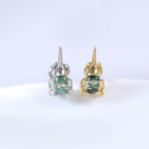 Könnte beinhalten: Ein Paar insektenförmige Ohrringe. Einer ist silberfarben, der andere goldfarben. Beide haben einen grünen, gesprenkelten Steinkörper. Die Ohrringe sind vor einem weißen Hintergrund platziert.