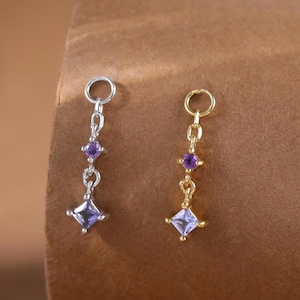 May include: Two dangling charms, one silver-toned and one gold-toned, each featuring a small round purple stone above a larger square-cut light purple stone. Both charms include a circular jump ring at the top for attachment to jewelry.