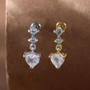 May include: A pair of earrings, one silver and one gold, each featuring a heart-shaped gemstone pendant. The earrings have a delicate design with small, light blue accent stones. The background is a warm, neutral tone.