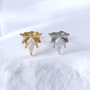 Puede incluir: Dos pendientes de bot&oacute;n, uno dorado y otro plateado, cada uno con una parte superior en forma de lazo y una gema transparente en forma de l&aacute;grima. Los pendientes se muestran sobre una superficie blanca texturizada.