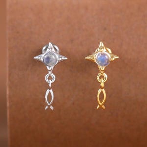 May include: A pair of stud earrings, one silver and one gold, each featuring a four-pointed star design with a small, round, blue gemstone at the center. Below the star, a delicate chain with a small, fish-shaped charm hangs.