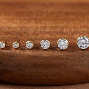 May include: A row of six round stud earrings in graduated sizes, each featuring a clear, faceted stone set in a gold-toned bezel. The earrings are arranged in a line from smallest to largest, showcasing a simple, circular design with metallic edges.