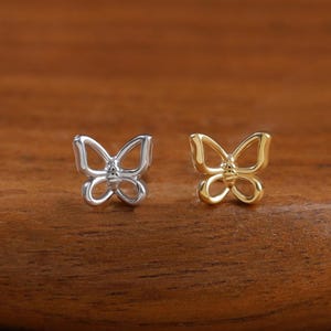 Puede incluir: Dos pequeños pendientes de botón con forma de mariposa. Un pendiente es de color plateado con un acabado metálico pulido, y el otro es de color dorado con un acabado metálico pulido a juego. Ambos presentan un diseño de alas caladas.