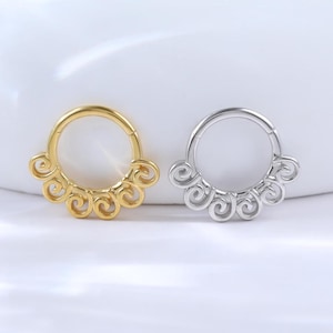 Pode incluir: Dois piercings decorativos para o nariz. Um é dourado, o outro prateado. Cada piercing tem um design circular com redemoinhos decorativos na borda. Os piercings são exibidos em uma superfície branca.
