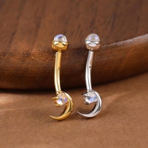 Può includere: Due piercing per l'ombelico, uno dorato e uno argentato, ciascuno con una barra curva, un ciondolo a forma di luna e una piccola gemma rotonda. I gioielli sono esposti su uno sfondo marrone caldo.
