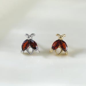 May include: Two stud earrings shaped like ladybirds, one in silver-tone metal and one in gold-tone metal. Each earring features two marquise-cut deep red gemstones forming the wings and a small metal bead representing the head of the insect.