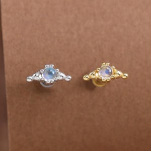 以下が含まれることがあります： シルバーとゴールドの2つのスタッドピアス。中央に丸い虹色の石が配置され、その周囲を小さな金属製のビーズと尖った三角形のフレームが囲んでいます。裏側には平らなポストが付いています。