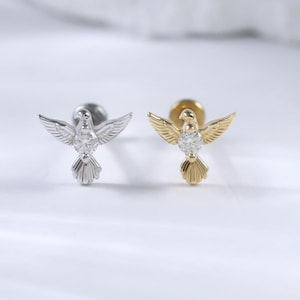May include: Two stud earrings, one silver and one gold, each shaped like a dove with outstretched wings. The doves have a small, clear gemstone on their chest. The earrings are on a white surface.