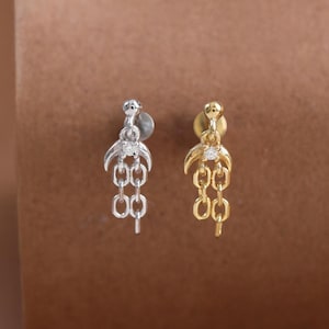 Pode incluir: Dois brincos de pino, um prateado e outro dourado, cada um apresentando uma forma de meia-lua no topo com uma pequena pedra transparente e dois elos de corrente curtos pendurados verticalmente a partir da base da lua.