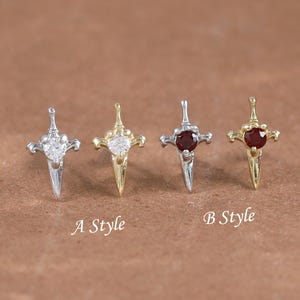Op de afbeelding: Vier kleine kruisvormige oorbellen. Twee zijn zilverkleurig met heldere stenen en twee zijn goudkleurig met rode stenen. De tekst "A Style" en "B Style" staat onder de oorbellen. Op een bruin oppervlak.