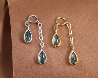 May include: Two pairs of earrings. One pair is silver-toned, the other is gold-toned. Each earring features a teardrop-shaped green gemstone suspended from a chain. The earrings are displayed on a brown surface.