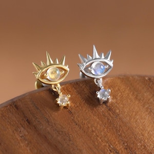 May include: Two earrings, one gold and one silver, each featuring an eye design with a crown and a dangling gemstone. The gold earring has a yellow gemstone, while the silver earring has a blue-white gemstone. The earrings are set against a wooden surface.