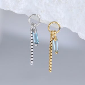 May include: Two earrings, one silver and one gold, each with a small hoop and dangling chain. Each earring features a light blue rectangular gem. The silver earring has a chain with square links, the gold with rectangular links.