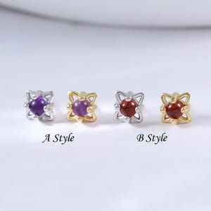 Può includere: Quattro piccoli orecchini a bottone con centri di pietre preziose. Due paia sono etichettati "A Style" e "B Style". Gli orecchini "A Style" hanno montature viola e oro, mentre gli orecchini "B Style" hanno montature rosse e oro. Gli orecchini sono su una superficie bianca.