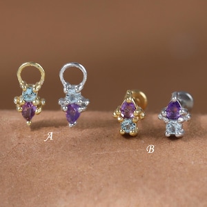 May include: Close-up of four small earrings. Two are gold-toned, and two are silver-toned. Each earring features a teardrop-shaped purple gemstone, surrounded by smaller blue and clear stones. The earrings are displayed on a brown surface.