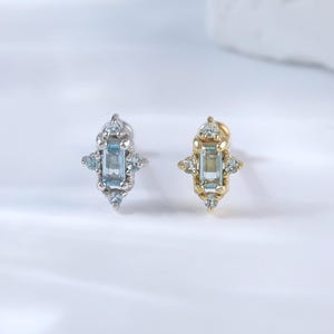 May include: Two stud earrings, one silver and one gold, each featuring a rectangular, light blue gemstone at the center. The earrings have a symmetrical design with smaller, clear gemstones surrounding the central stone, creating a sparkling effect.