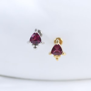 May include: Two stud earrings featuring triangular, deep red gemstones. One earring has a silver-coloured post and setting, the other a gold-coloured post and setting. The earrings are displayed on a white surface.