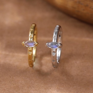 May include: Two small hoop earrings, one gold and one silver, each featuring a small, oval-shaped, iridescent stone. The earrings are set against a warm brown background, with a wooden element in the upper right corner.