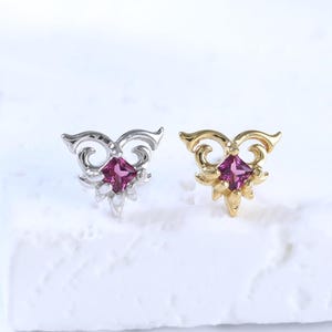 Peut inclure: Deux boucles d'oreilles à tige avec une pierre précieuse magenta de taille carrée sertie dans une monture en métal décorative. L'une est argentée et l'autre dorée. Les deux présentent un motif complexe et symétrique entourant la pierre.