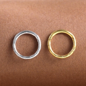 Può includere: Due piccoli anelli strutturati. Uno è color argento, l'altro dorato. Gli anelli sono circolari e hanno un aspetto martellato o strutturato. Lo sfondo è di colore marrone pieno.