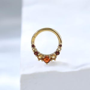 May include: A gold hinged hoop ring featuring a cluster of small, faceted gemstones in warm tones of orange, yellow, and deep red. The stones are arranged in a graduated arc along the lower portion of the circular metal band, creating a decorative accent.