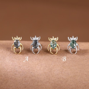 Könnte beinhalten: Vier insektenförmige Ohrstecker in einer Reihe angeordnet. Zwei Ohrringe sind goldfarben und zwei sind silberfarben, wobei jeder in der Mitte einen ovalen grünen Edelstein aufweist. Die Buchstaben A und B sind unter den Ohrringen abgedruckt.