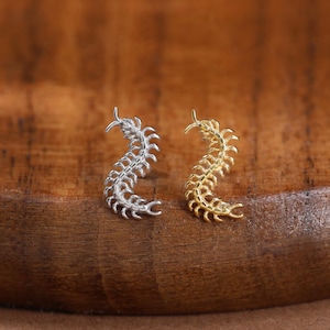 May include: Two small stud earrings shaped like centipedes, one finished in a bright silver tone and the other in a warm gold tone. Each earring features a curved, segmented body with multiple tiny legs extending from the sides in a detailed design.