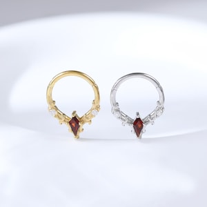 May include: Two small, decorative rings. One is gold-toned, the other silver-toned. Each ring features a central, teardrop-shaped, red gemstone and smaller white gemstones. The rings are displayed on a white, curved surface.