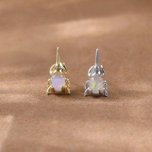 Könnte beinhalten: Ein Paar kleiner Ohrstecker. Einer ist goldfarben, der andere silberfarben. Jeder hat einen kleinen, hellfarbigen Opalstein. Die Ohrringe sind vor einem warmbraunen Hintergrund platziert.