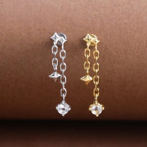 Peut inclure: Deux boucles d'oreilles pendantes, l'une argentée et l'autre dorée, chacune ornée d'un clou en forme d'étoile, d'une chaîne, d'un petit cône et d'une pierre précieuse ronde et transparente. Fond marron.
