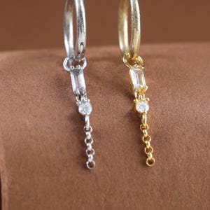 Puede incluir: Dos pendientes de aro, uno plateado y otro dorado, cada uno con un colgante compuesto por una pequeña piedra rectangular, una piedra redonda diminuta y un delicado segmento de cadena que cuelga desde la base del aro.