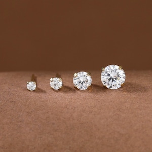 May include: Four round stud earrings arranged in a row by size, from smallest to largest. Each earring features a single clear, faceted stone set in a gold-toned metal prong setting, showcasing a brilliant sparkle and a classic, simple design.