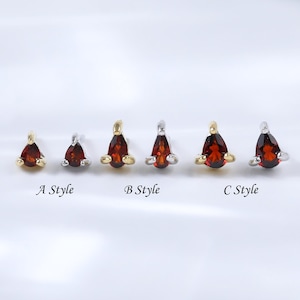 Pode incluir: Uma coleção de pequenos brincos de pressão com pedras preciosas vermelhas em forma de gota. Os brincos são cravejados em metal dourado e prateado, com três estilos diferentes rotulados A, B e C. Os brincos são exibidos em uma superfície branca.