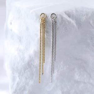 May include: Two pairs of drop earrings, one gold and one silver, presented on a white, textured block. The earrings feature a fine chain design, with a small loop at the top for fastening.