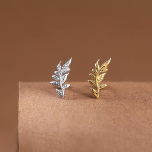 May include: Two stud earrings shaped like delicate leaves, one in a polished silver tone and the other in a bright gold tone. Each earring features a slender stem with small, pointed leaf segments extending from the centre, creating a symmetrical botanical design.