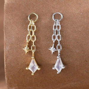 May include: Two dangle earrings, one gold-toned and one silver-toned, each with a chain, a star charm, and a diamond-shaped, faceted gemstone. The earrings are set against a brown background.