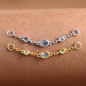 Könnte beinhalten: Zwei filigrane Kettenverbinder, einer silberfarben und einer goldfarben, jeweils mit kleinen, hellblauen ovalen und runden Edelsteinen verziert. Die Ketten werden auf einer braunen Oberfläche präsentiert, die das Design des Schmucks hervorhebt.
