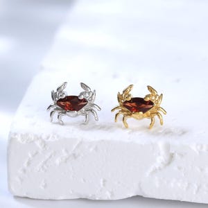May include: A pair of stud earrings shaped like crabs. One earring is silver-toned and the other is gold-toned. Each crab features a dark red, faceted gemstone set in the center of its body, with detailed legs and claws extending outward.