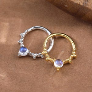 Puede incluir: Dos pendientes de aro, uno plateado y otro dorado, cada uno con una pequeña gema redonda de color azul claro. El pendiente plateado tiene un diseño de cuentas, mientras que el pendiente dorado tiene un patrón texturizado. Los pendientes están sobre una superficie marrón.