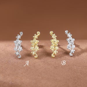 以下が含まれることがあります： 小さな透明な石をあしらった花のつるのデザインが特徴の、カーブしたスタッドピアス4つが並んでいます。2つはシルバーカラー、2つはゴールドカラーです。ピアスの下には、2つのセットを示す文字AとBが印刷されています。