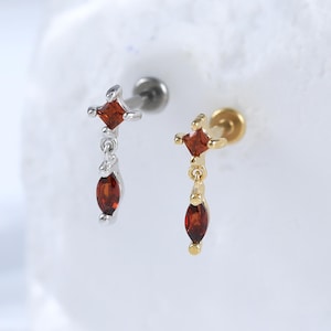 May include: Two earrings with a silver and gold finish. Each earring features a small, square, red gemstone at the top, with a dangling, oval-shaped red gemstone below. The earrings are displayed on a white surface.